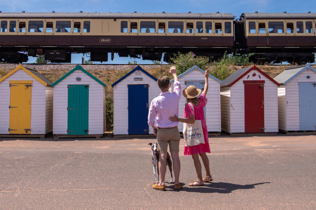 Couple with dog waving at steam train in front of beach huts at Goodrington